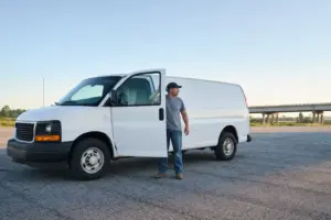 A cargo van owner-operator standing at the open driver door of a white cargo van on an empty lot at sunrise